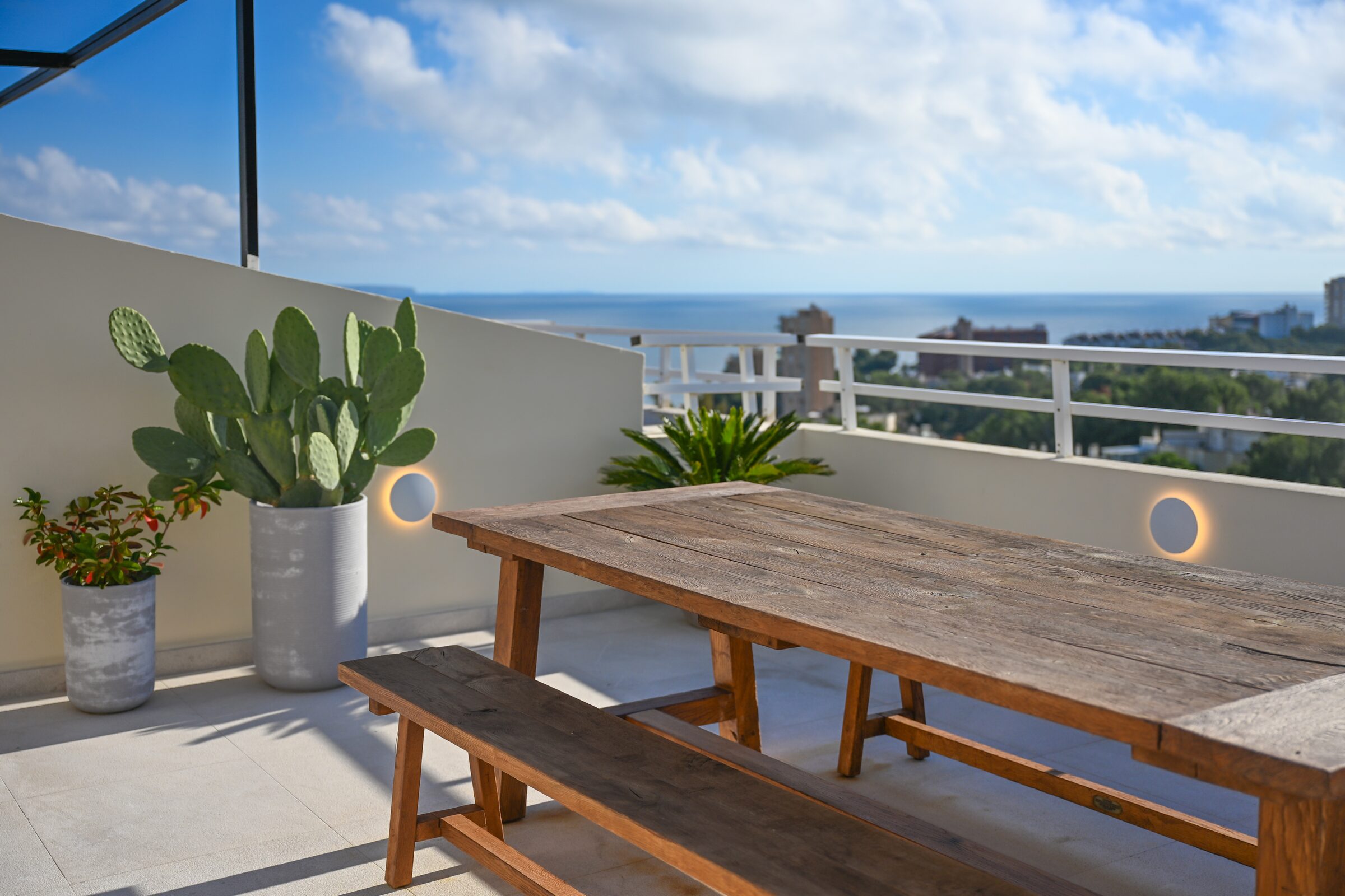 Rooftop terrace of Cas Blau in Cas Català, Mallorca, with a long reclaimed-wood bench-table, cactus and the Bay of Palma under a wide cloud-streaked sky.