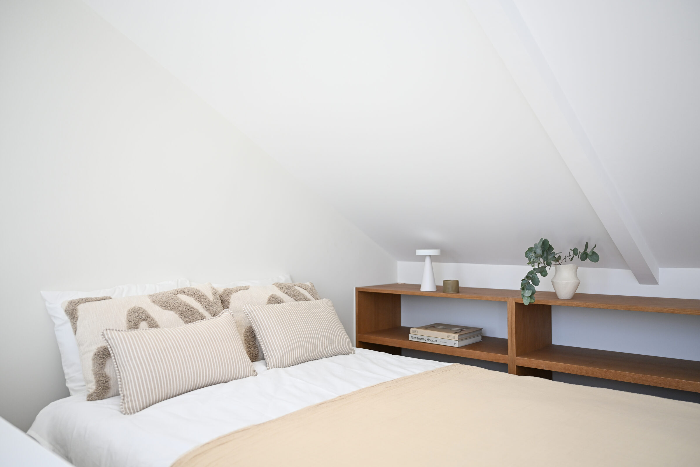 Side view of the bedroom with patterned linen pillows and a long low oak shelf running under the eaves.