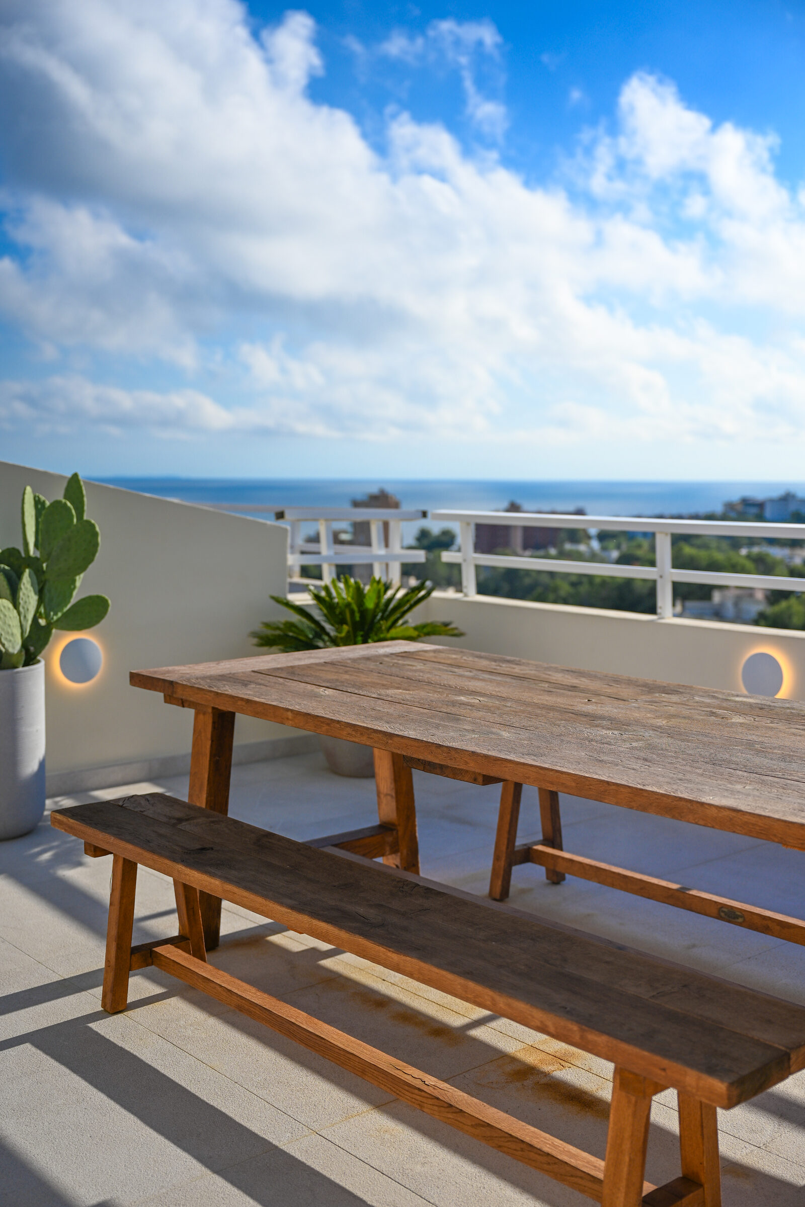 Long reclaimed-wood bench-table on the rooftop in full daylight, framed by cactus and the Bay of Palma.