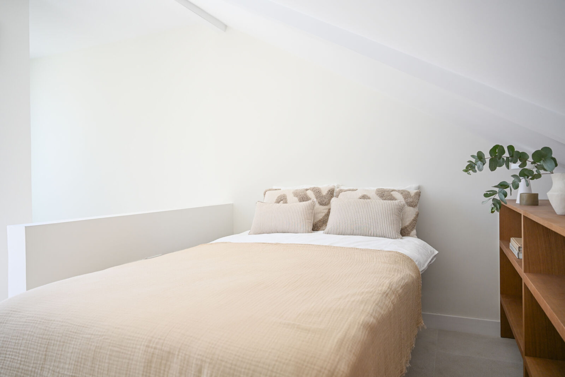 Bedroom under the sloped ceiling with caramel bedding, patterned linen pillows and an oak shelf with eucalyptus.