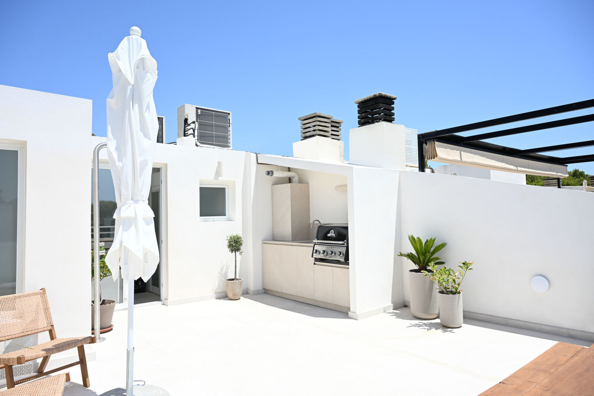 Wide angle of the rooftop terrace with cane chair, closed parasol, outdoor kitchen with grill and steel pergola against the sky.