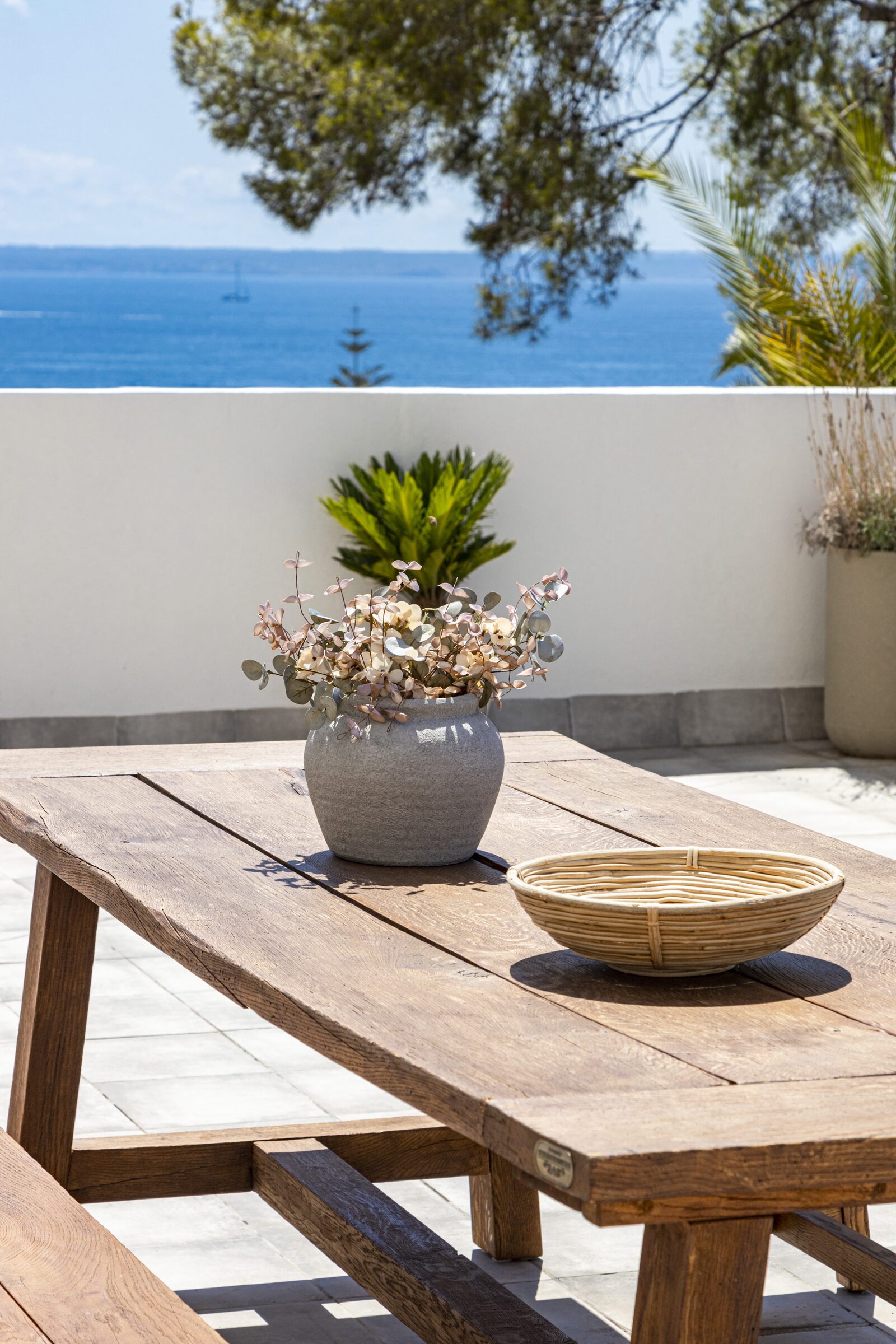 Outdoor table styled with a stoneware planter and rattan bowl, with a glimpse of the bay through the pines.