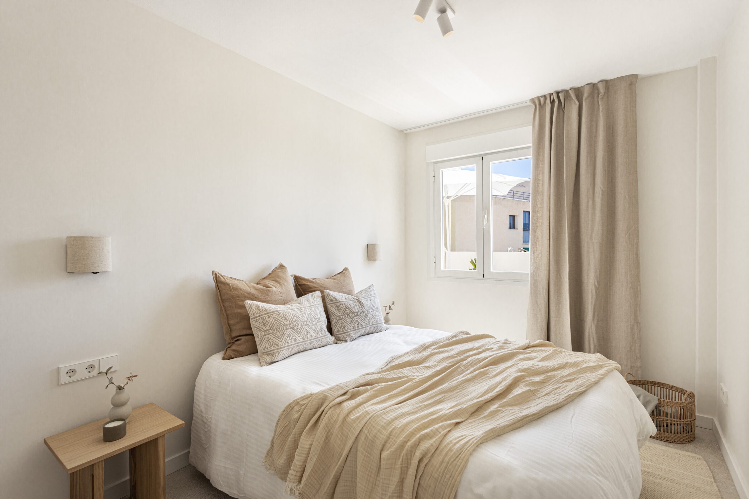 Calm bedroom with caramel linen throw, soft pillows and a wooden side table beneath the window.